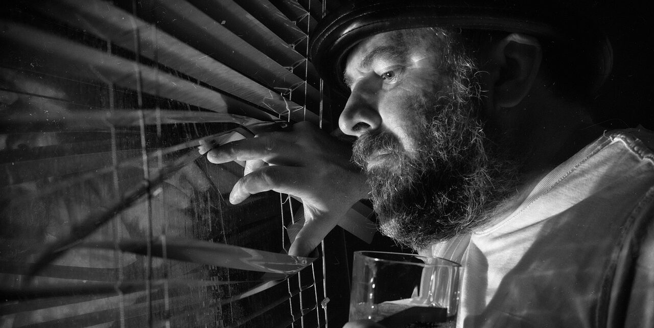 “Man looking out window through blinds with cautious expression, symbolizing the scanning and fear experienced in social anxiety.”